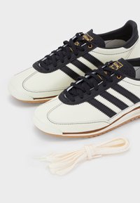 Een paar witte en zwarte Adidas sneakers met zwarte veters en een extra set witte veters op een effen achtergrond.