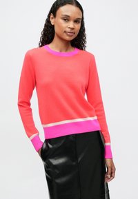 Mujer vestida con un suéter de color coral con ribetes en rosa brillante y beige, combinado con una falda de cuero negra, de pie con las manos en los bolsillos.