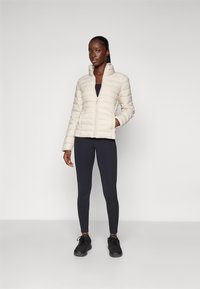 Lichtbeige pufferjack met verticale quilt, ritsluiting en hoge kraag, gecombineerd met zwarte leggings en zwarte sneakers.