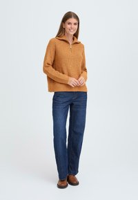 Rostfärgad stickad tröja med dragkedja i kragen, i kombination med mörka denimjeans och bruna ankelstövlar. Texturerat tyg, avslappnad passform.