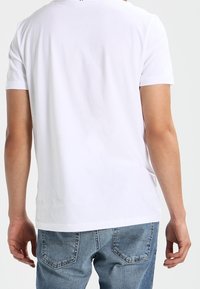 Aikuinen, joka käyttää yksinkertaista valkoista lyhythihaista t-paitaa ja sinisiä farkkuja, näkyy selkäpuolelta tasaisen taustan edessä.