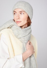 Heledhall hallide ribakudumine beanie ja vastava lapiga pehme materjali, vabalt kaela ümber rippuv, kombineeritud koorevalge fliisvestiga.