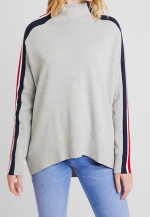 Kvinde iført en lysegrå sweater med marineblå, rød og hvid striber ned langs ærmerne og blå jeans, stående foran en ensfarvet baggrund.