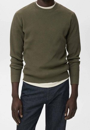 Homme portant un pull côtelé vert olive par-dessus une chemise blanche avec un pantalon bleu foncé sur un fond uni.