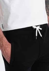 Pantaloni jogger neri con vita regolabile e tasche laterali. La cintura presenta una texture a coste. Un'etichetta bianca è visibile sul lato.