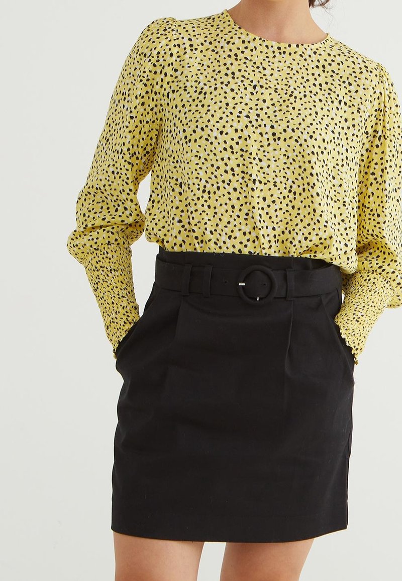 Blusa gialla con motivo a pois neri, maniche lunghe, scollatura rotonda. Abbinata a una gonna nera a vita alta con una cintura dotata di fibbia circolare.