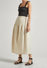 Mustassa rypistetty crop-top, jossa on frillaolkapäät, yhdistetty beige raidalliseen kietaisuhameeseen. Malli käyttää mustia litteitä sandaaleja, joissa on soljet.