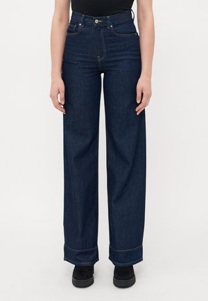 Kvinne iført høyt liv, mørkeblå vide jeans med oppbrettede hems og svarte støvler, stående mot en enkel lys bakgrunn.