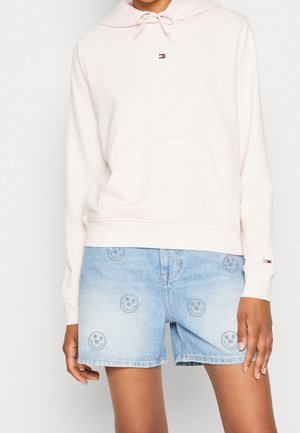 Person iført lyserød hættetrøje med frontlomme og lyseblå denimshorts med små smiley-ansigtsprint.