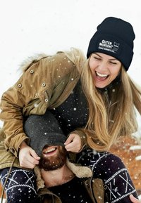 Black knitted beanie with white text and owl graphic, worn by a person with long hair, outdoors in the snow, paired with a brown coat.