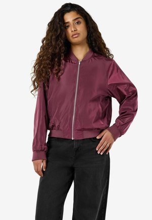 Femme aux longs cheveux bouclés portant une veste bomber bordeaux zippée et un pantalon noir, debout avec une main sur la hanche, sur fond blanc.