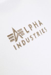 Tessuto bianco con logo ricamato beige che presenta il testo "ALPHA INDUSTRIES" e un design stilizzato a stella.