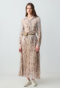 Lungo vestito trasparente con stampa di serpente in tonalità beige e marrone, caratterizzato da un colletto e chiusura con bottoni. Indossato con una cintura dorata e sneakers bianche.