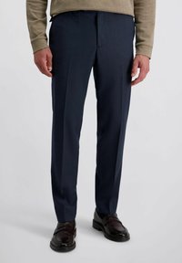 Man in een donkerblauwe pantalon, bruine leren instappers en een beige gestreept overhemd met lange mouwen, staande op een lichtgrijze achtergrond.