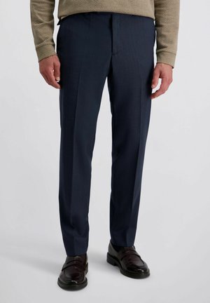 Man in een donkerblauwe pantalon, bruine leren instappers en een beige gestreept overhemd met lange mouwen, staande op een lichtgrijze achtergrond.