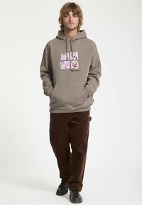 Brown sweatshirt with graphic print featuring faces and text, paired with brown corduroy pants and black shoes. Soft texture, relaxed fit.