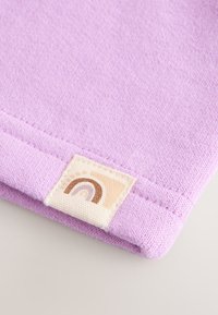 Lähikuva taitetusta laventelinsävyisestä kankaasta, jossa on ommeltu helmava on ja pieni beige kyltti, jossa on minimalistinen ruskea sateenkaarisymboli.