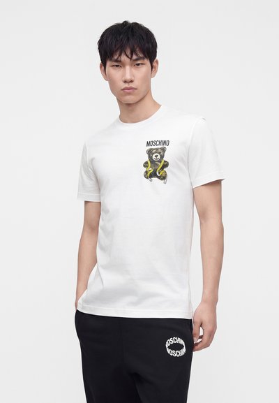 Νέος άνδρας με λευκό μπλουζάκι Moschino με σχέδιο αρκουδάκι και μαύρο παντελόνι Moschino, στέκεται μπροστά σε απλό φωτεινό φόντο.
