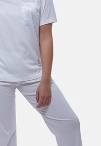 Henkilö, joka on pukeutunut valkoiseen lyhythihaiseen paitaan, jossa on satiininen rintatasku, ja valkoisiin housuihin. Hän seisoo toinen jalka koukistettuna ja käsi rentona.