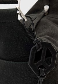 Primo piano di una borsa in pelle nera con texture, dotata di una chiusura metallica impressa con la scritta "ALLSAINTS" e un simbolo di pace in pelle attaccato da un cordino annodato.