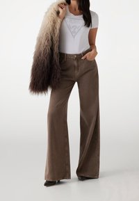 Pantalones vaqueros marrones de pierna ancha combinados con una camiseta blanca con un gráfico plateado, acentuados con una prenda exterior de piel sintética texturizada con colores degradados.