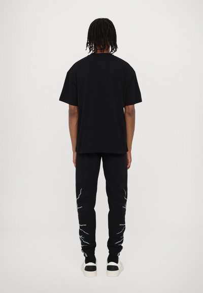 T-shirt oversize nera indossata con pantaloni da tuta neri caratterizzati da disegni a forma di fulmine bianchi e sneakers in bianco e nero. Vista da dietro.