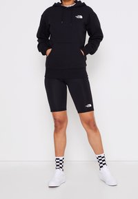 Černá mikina s kapucí a přední kapsou s logem, doplněná černými cyklistickými šortkami. Bílá teniska a kostkované ponožky dokončují sportovní vzhled.