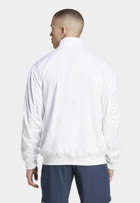Witte lichte jas met hoge kraag, elastische manchetten en zoom. Glad textuur met minimale naaddetails, gedragen over marineblauwe shorts.