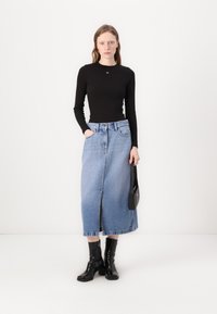 Svart langermet topp, blå denim midi-skjørt med frynser i kantene, og svarte hæler. Modellen holder en liten svart håndveske.