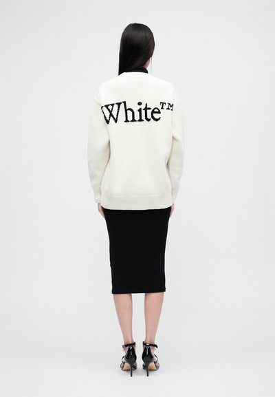Κρέμ oversize πουλόβερ με το "White™" σε μαύρη πλέξη στη πλάτη, συνδυασμένο με μια εφαρμοστή μαύρη φούστα και μαύρες ψηλές γόβες με λουράκια.