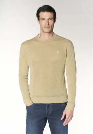 Uomo che indossa un maglione beige a maniche lunghe con piccolo logo giallo e jeans blu, in piedi con una mano in tasca su sfondo bianco.