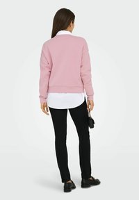 Vrouw met lang bruin haar, een roze sweatshirt over een wit shirt, zwarte broek, zwarte schoenen, die een kleine zwarte handtas vasthoudt en staat.