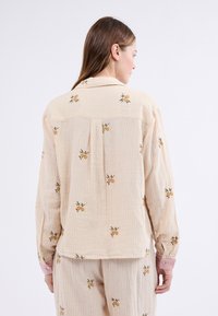 Camisa a rayas beige con bordado floral en amarillo y verde, con puños abotonados y un corte relajado; presenta un detalle de pliegue en la parte trasera.
