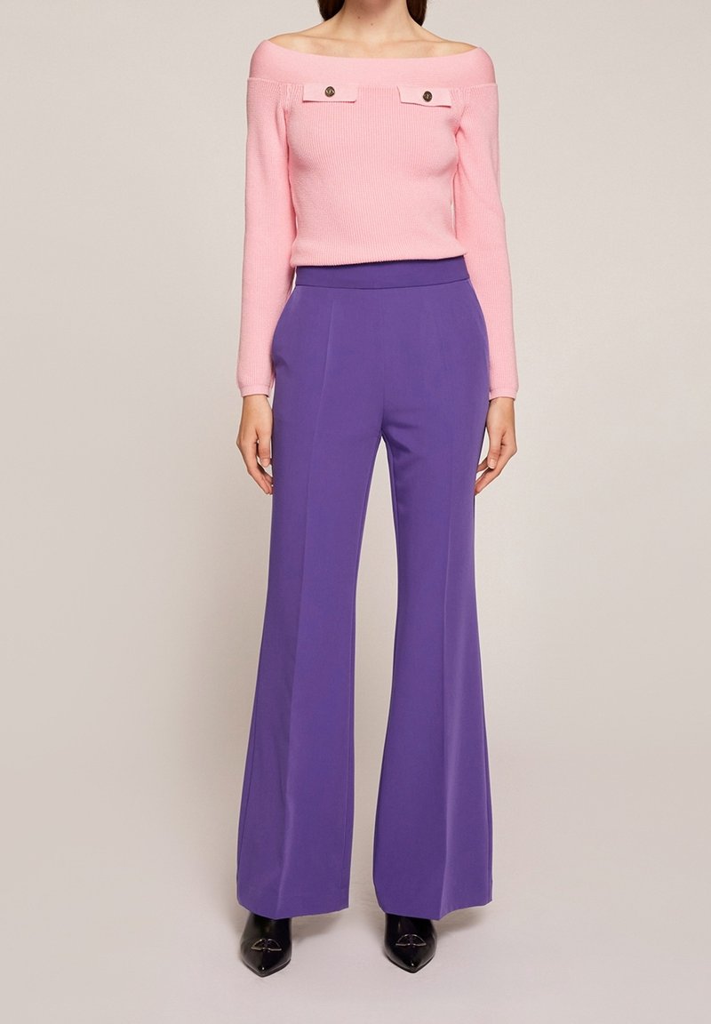 Kvinna klädd i en rosa off-shoulder ribbad tröja med knappdekorer och högmidjade lila vida byxor med svarta spetsiga skor.