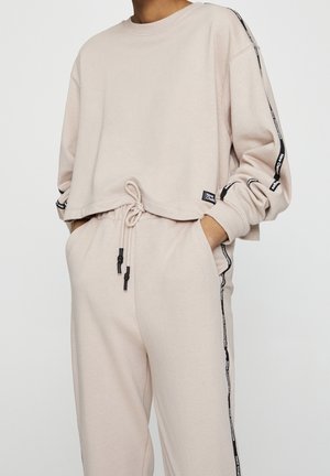 Personne portant un vêtement d’intérieur beige avec des cordons noirs et des bandes latérales noires étiquetées "New Classic" sur les manches et le pantalon, mains dans les poches.