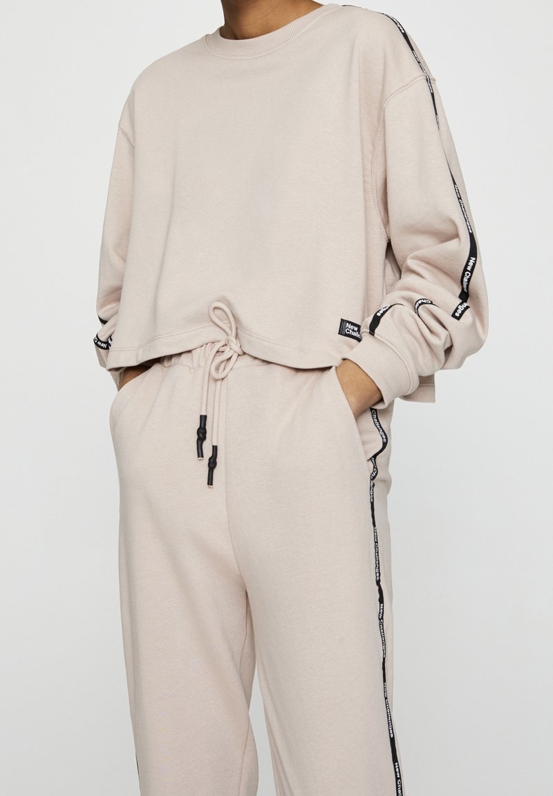 Personne portant un vêtement d’intérieur beige avec des cordons noirs et des bandes latérales noires étiquetées "New Classic" sur les manches et le pantalon, mains dans les poches.