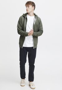 Groene rits hoodie met zijzakken, gedragen over een wit T-shirt, gecombineerd met donkere broek en witte sneakers met bruine accenten.