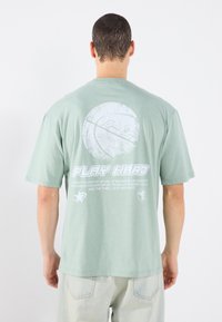 Gaiši zaļš pārmēru t-krekls ar basketbola grafiku un tekstu "SPĒLĒ HARD" baltā krāsā uz muguras. Kokvilnas materiāls, īsas piedurknes.