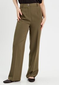 Pantalons large en vert olive fabriqués en tissu lisse, dotés d'une fermeture à bouton, de passants de ceinture et de poches avant. Portés avec des ballerines noires.