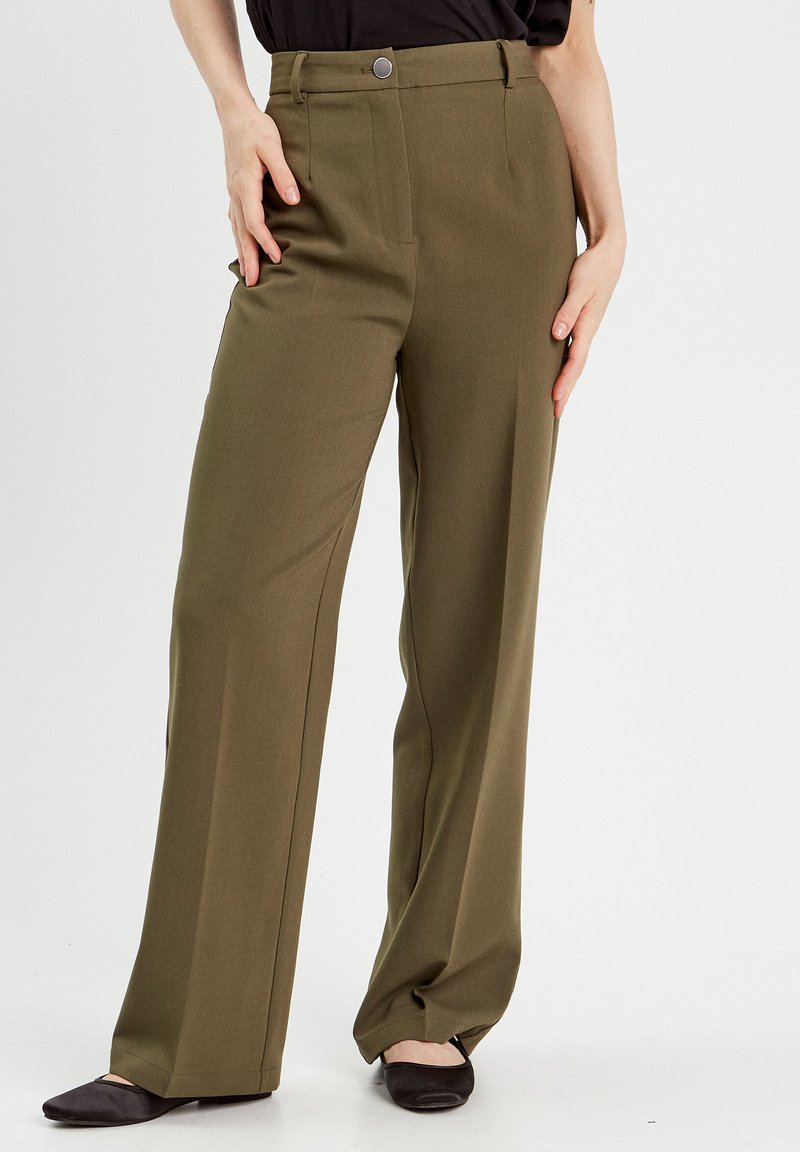 Pantalons large en vert olive fabriqués en tissu lisse, dotés d'une fermeture à bouton, de passants de ceinture et de poches avant. Portés avec des ballerines noires.