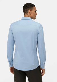 Camicia azzurra a maniche lunghe con colletto a punta, texture liscia, polsini con bottone e orlo curvo, vista da dietro.