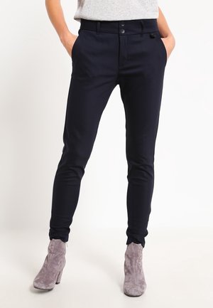 Vrouw die zwarte slim-fit broek draagt met handen in de zakken en grijze suède enkellaarsjes, staand tegen een witte achtergrond.