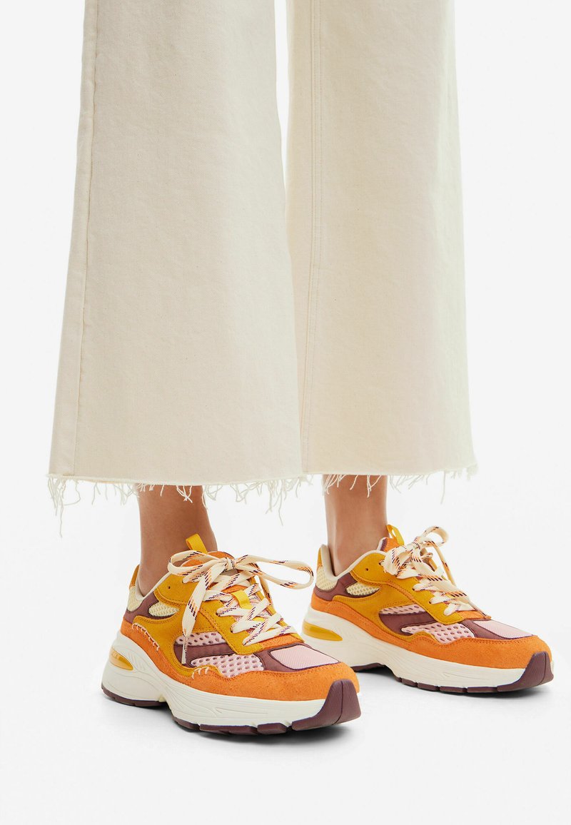 Las zapatillas coloridas presentan una mezcla de materiales en naranja, amarillo y marrón. El diseño incluye acentos de malla y suelas gruesas, con cierres de cordones.