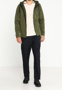 Chaqueta de forro polar verde con cremallera y capucha, llevada sobre una camisa clara; combinada con pantalones negros y zapatillas deportivas negras.