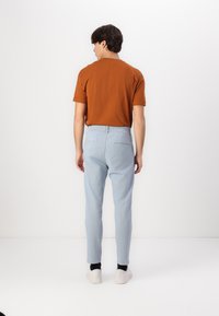 Pantalon bleu clair avec taille élastique et poches arrières, associé à une chemise marron à manches courtes. Modèle tourné de dos par rapport à l'appareil photo.