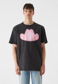 Tumši pelēks kokvilnas T-krekls ar rozā kovboju cepures grafiku un baltiem burtiem. Brīvs piegriezums, īsas piedurknes un apaļa kakla izgriezums.