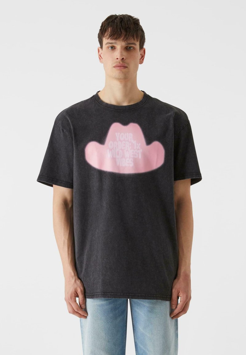 Tumši pelēks kokvilnas T-krekls ar rozā kovboju cepures grafiku un baltiem burtiem. Brīvs piegriezums, īsas piedurknes un apaļa kakla izgriezums.