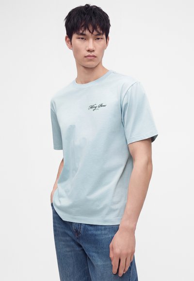 Νέος άνδρας με ανοιχτό γαλάζιο γραφικό t-shirt και μπλε τζιν, στέκεται μπροστά σε απλό λευκό φόντο.
