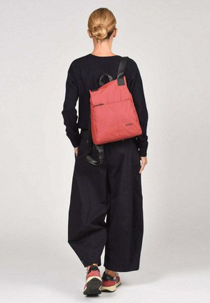 Mochila de nylon roja con un diseño elegante y sencillo, que cuenta con una asa superior, una correa negra ajustable y un bolsillo frontal. Logotipo de la marca visible.