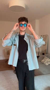 Zilā un baltā svītraina pārāka krekls, melns T-krekls zem tā, melnas bikses un zilas saulesbrilles ar atstarojošām lēcām.